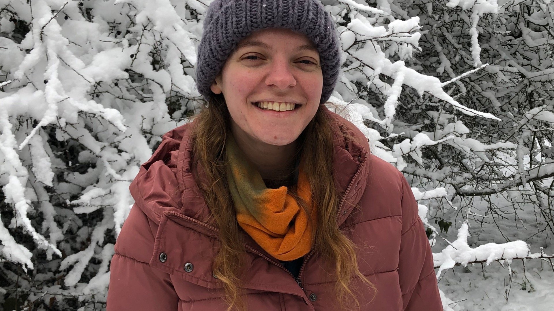 Emily, a smiling caucasian woman with brown hair standing outside in the snow wearing a hat and scarf