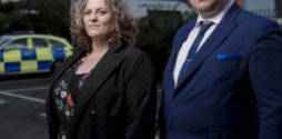 A male and female lawyer stand in a police station car park. Both are looking powerfully at the camera.