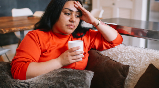 A young woman looks off to the side while sat on a sofa holding a cup.