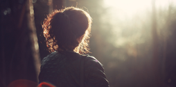 A woman with her back to us seems to be looking out into the distance. She appears to be in woodland.