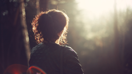 A woman with her back to us seems to be looking out into the distance. She appears to be in woodland.