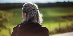 A young woman with short silver hair stands outside looking at a field. The photo is taken from the back so we can't see her face.