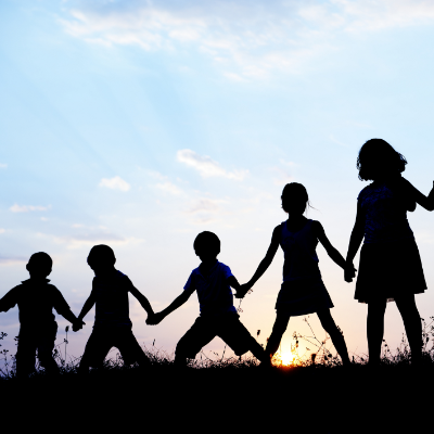 A group young children stand in a line while holding hands and appearing to be playing. They have their backs to us and are silhouetted against the sky.