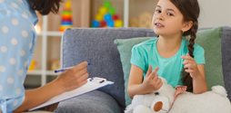 A young girl with pigtails sits on a sofa, holding a stuffed toy and talking to a woman counsellor who is sat on a chair in front of her.