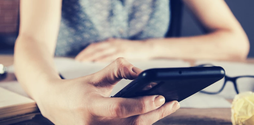 A woman sits at a table using her smartphone, surrounded by a notebook and a pair of glasses. The woman's face is not in the frame.