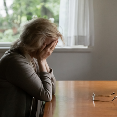 An older woman sits at a table with her head in her hands. Her glasses sit on the table in front of her.