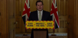 MP Robert Jenrick stands at a podium with 'Stay home, protect the NHS, save lives' on it. Behind him are to Union Jack flags.