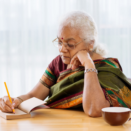 An elderly woman sits at a table and is writing in her journal.
