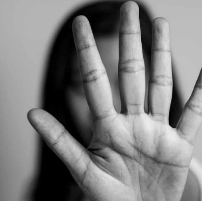 A black and white image of a woman standing opposite a camera.  Her hand is held up in a stop sign, hiding her face.