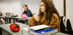 Side shot of a young woman sitting at a desk in school looking ahead of her. Her pencil case and two academic books are also on the desk.