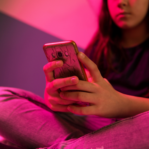 A teenage girl looks at a mobile phone while slumped against a wall. She has a serious expression on her face.