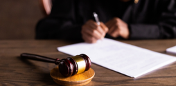 A wooden gavel rests on top of a wooden desk. In the background, out of focus is someone writing on a note pad- we can't see their face.