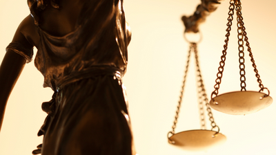 The image shows a photo of the back of a statue of Lady Justice, holding up scales in her right hand.