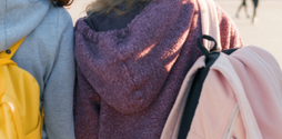 The photo shows two girls with their backs to us, standing in a square. Both have backpacks on.