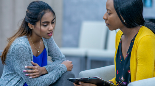 A female counsellor holding a clipboard and her female patient sit on armchairs. The female patient is mid speech and looking at the clipboard and the counsellor is looking at her.