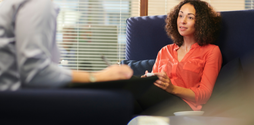A woman in her 30s or 40s with brown curly hair and wearing an orange shirt sits in a blue chair facing her therapist in the therapist's office.