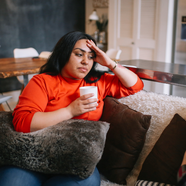 A young woman with long black hair sits on a sofa while holding a mug. She has one elbow resting on the back of the sofa as she presses the hand on her forehead.