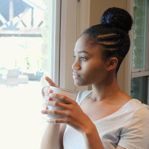 A young woman stands inside a house holding a hot drink in a mug and looking off to the side. Her expression is serious.