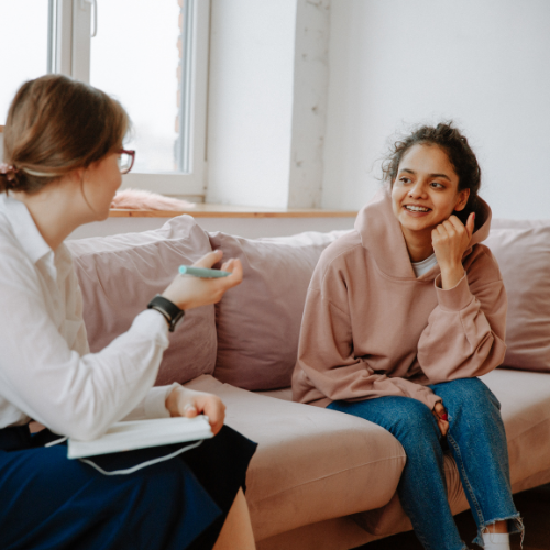 A young woman wearing a beige hoodie and blue jeans talks to a young female counsellor. Both are sitting on a sofa.