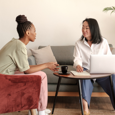 A young woman sits in a chair while talking to an older woman who is a counsellor. The older woman is sat on a sofa next to the chair and is using a laptop on a table.